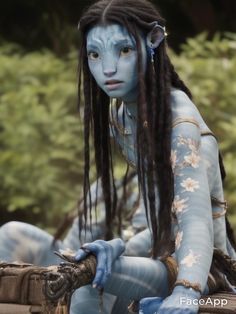 a woman with blue paint on her body and long dreadlocks sitting in front of trees