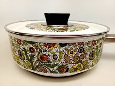 an ornate pot with black lid on a white tableclothed surface, decorated with colorful flowers and leaves