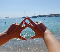 two hands making the shape of a heart with boats in the water and people swimming in the background