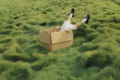 a man laying in a cardboard box on top of a green grass covered field with his feet up