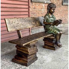 a statue sitting on top of a wooden bench next to a brick building and holding a book