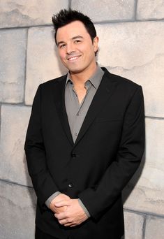 a man in a black suit and grey shirt smiling at the camera with his hands on his hips