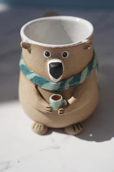 a ceramic bear with a scarf around its neck holding a cup in it's mouth