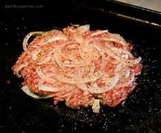 an uncooked hamburger with onions and seasoning on it in a frying pan