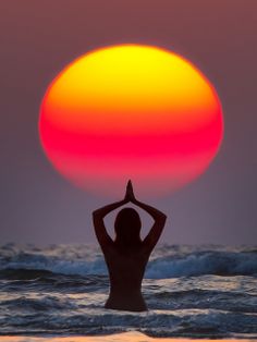 a woman standing in the ocean with her hands up to her head as the sun sets