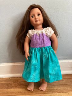 a doll with long brown hair wearing a blue and purple dress, standing against a wall