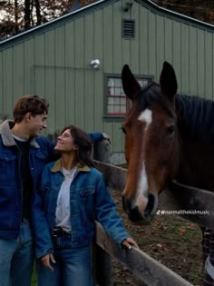 a man and woman standing next to a brown horse near a fence in front of a green building