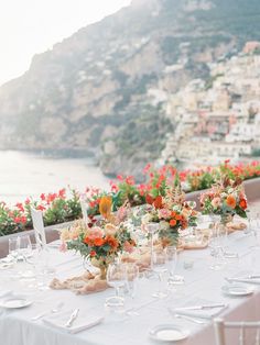 a long table with white linens and flowers on it is set for an outdoor dinner overlooking the ocean