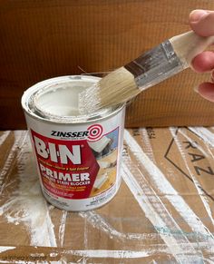 a person holding a paint brush over a can of white primer on top of cardboard