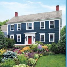 a blue house with white trim and red door surrounded by green grass, shrubs and flowers