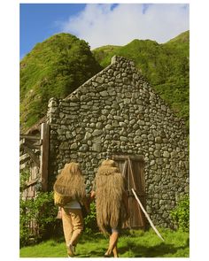 two people walking towards a stone building with grass on the ground and mountains in the background