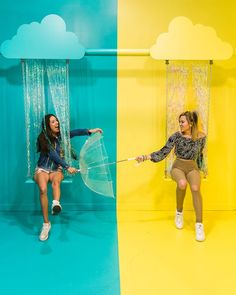 two women are posing in front of a backdrop with clouds and umbrellas on it