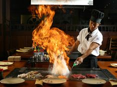 a chef is cooking on a grill with flames in the air and plates around him