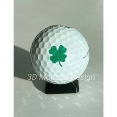 a white golf ball with a green shamrock on the front and bottom, sitting on a black stand