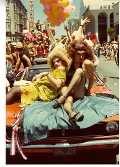 two women sitting on the hood of a car with balloons in the air above them