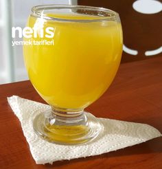 a glass filled with yellow liquid sitting on top of a wooden table next to a white napkin