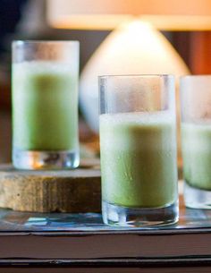 three glasses filled with green liquid sitting on top of a table next to a lamp