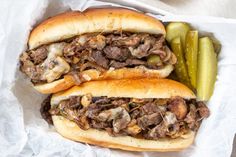 two sandwiches with meat, mushrooms and pickles in paper wrapper next to pickle