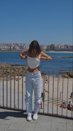 a woman in white shirt and jeans standing next to a metal fence near the beach