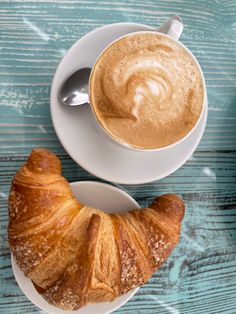 a croissant and cup of coffee on a blue wooden table with a spoon