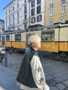 a woman with blonde hair standing in front of a yellow and white train on the street