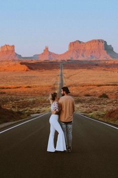 a man and woman walking down the middle of an empty road with mountains in the background