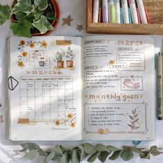 an open planner book sitting on top of a table next to some plants and markers