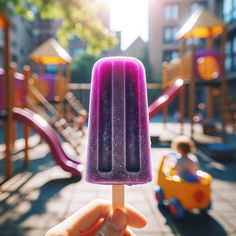 a hand holding an ice lollypop in front of a playground