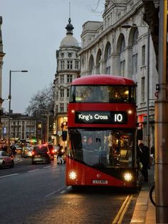 a red double decker bus driving down a street next to tall buildings and traffic lights