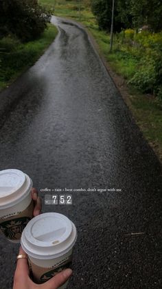 two cups of coffee are being held in the middle of a road with trees and bushes on both sides