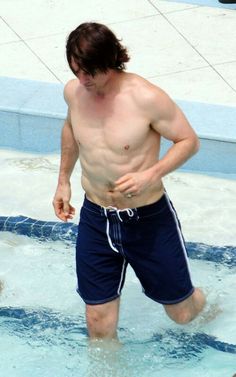 a shirtless man is walking in the water with his hand on his hip while wearing swimming trunks