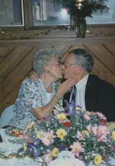 an older couple kissing each other while sitting at a table with flowers in front of them