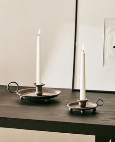 two white candles are sitting on a black table with silver plates and saucers in front of it