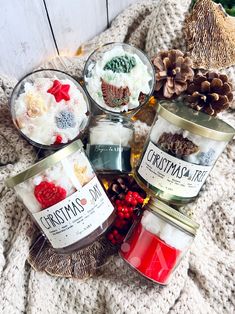 four jars filled with food sitting on top of a blanket next to pine cones and christmas decorations