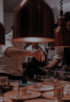 two chefs preparing food in a kitchen with plates on the table and lights hanging from the ceiling