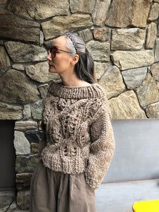 a woman standing in front of a stone wall wearing a brown sweater and tan pants