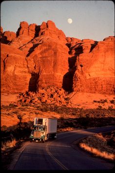 a truck is driving down the road in front of some red rocks and a full moon