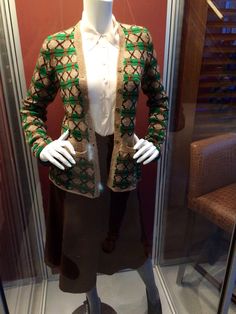 a mannequin wearing a green and gold jacket in a glass case with a brown chair behind it