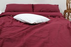 an unmade bed with red sheets and white pillow on the bottom, next to a wicker chair