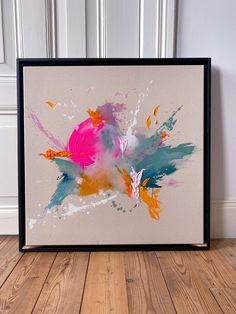 an abstract painting is displayed in a black frame on a wooden floor next to a white wall