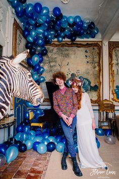 a man and woman standing next to a zebra in front of blue balloons on the wall