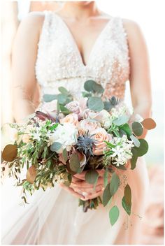 a bride holding a bouquet of flowers and greenery in her hands, wearing a wedding dress