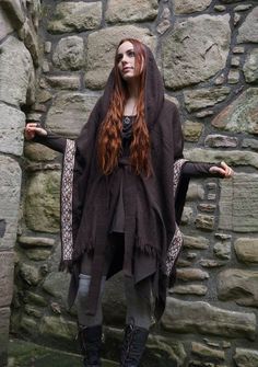 a woman with long red hair standing in front of a stone wall wearing boots and a shawl