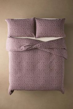 a bed with purple sheets and pillows on top of it, in front of a beige wall