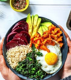 a person holding a bowl filled with vegetables and an egg on top of some rice