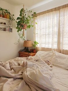 an unmade bed with white sheets and plants on the window sill next to it