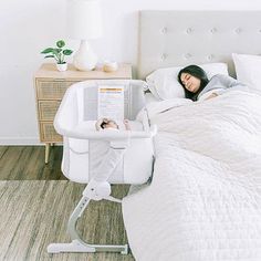 a woman laying in bed next to a white baby seat and table with a magazine on it