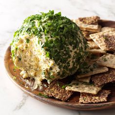 a wooden plate topped with crackers and a ball of cheese covered in green leafy toppings