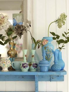 blue vases and flowers on a shelf in front of a white wall with a mirror