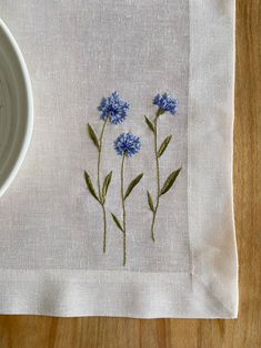 blue flowers are embroidered onto a white linen placemat on a wooden surface next to a bowl